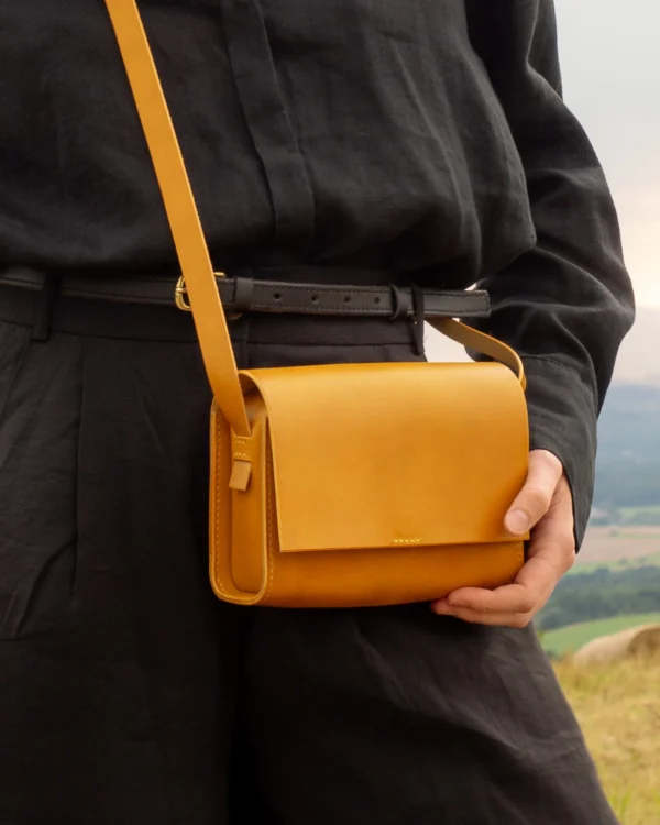 torebka-nr1-habaz-musztardowa-stylizacja-na-ramieniu Detal skórzanej torebki musztardowej Habaż na ramieniu modelki na tle wiejskiego krajobrazu.