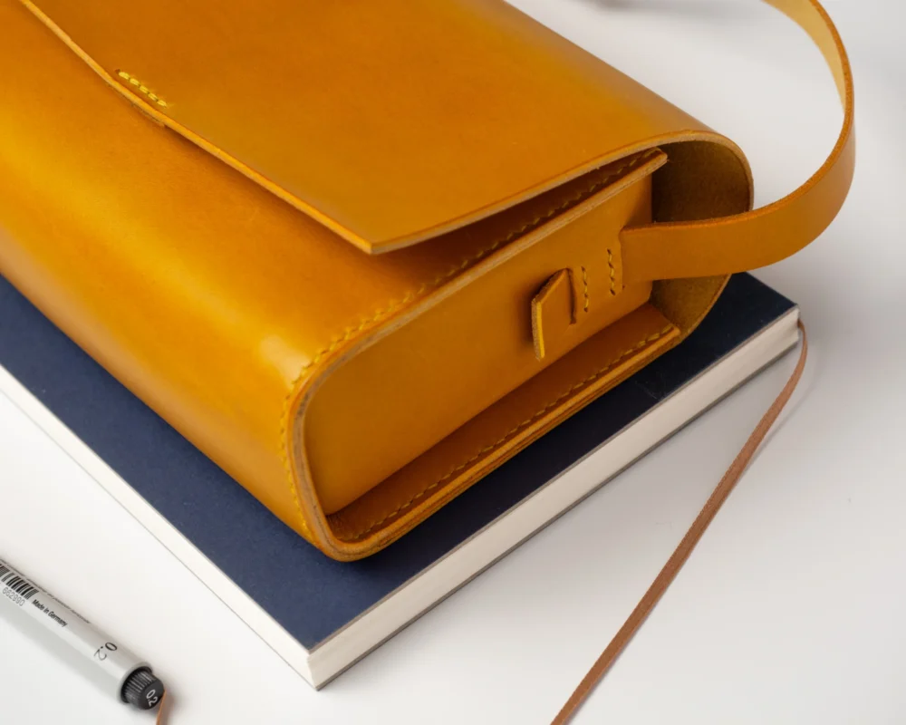 torebka-nr1-habaz-musztardowa-zbliżenie-na-notatniku Zbliżenie na musztardową torbę Habaż spoczywającą na niebieskim notesie i białym tle.