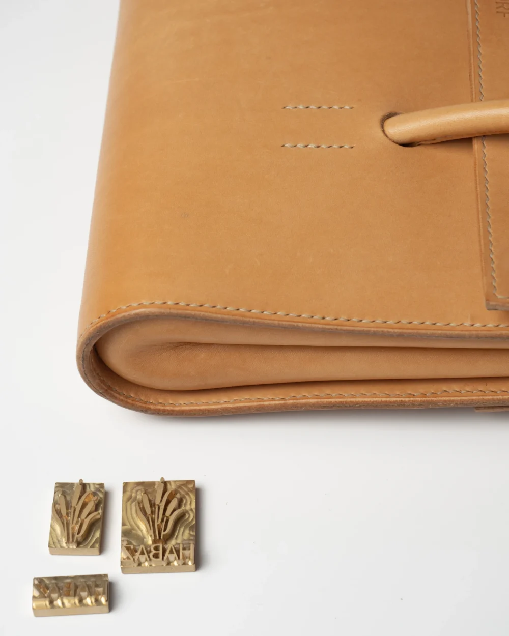 torebka-nr2-habaz-naturalna-detal-logo-stemple Zbliżenie na zaokrąglone rogi naturalnej torebki i obok metalowe stemple z logo Habaż.