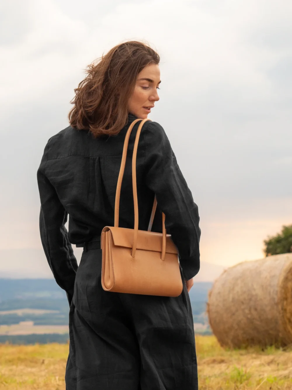 torebka-nr2-habaz-naturalna-stylizacja-z-na-tle-gor Modelka z naturalną torebką Habaż na ramieniu stoi na polu ze snopem siana w tle.