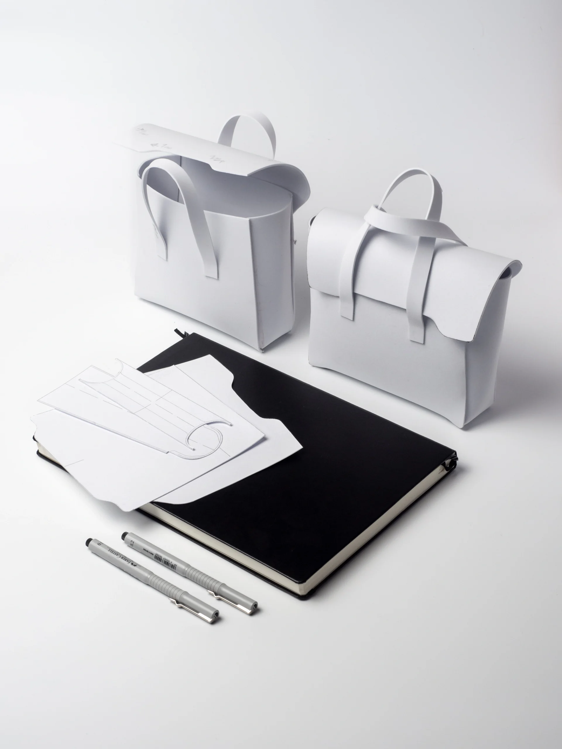 Proces projektowania torebek Habaź: dwa białe prototypy papierowe torebek, leżące obok czarnego szkicownika, rysunków kroju i dwóch cienkich pisaków.