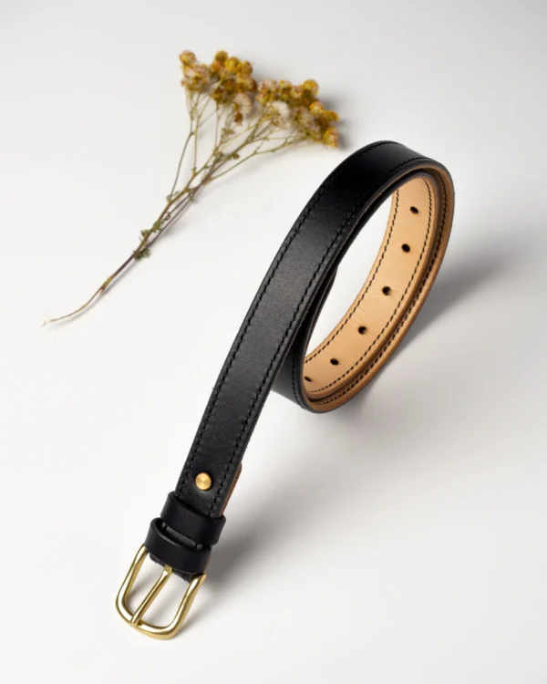 pasek-buttero-habaz-czarny-glowny-mosiadz Czarny, skórzany pasek Buttero, zwinięty, z mosiężną klamrą, obok suchych żółtych kwiatów. Widoczny naturalny, jasny spód paska.