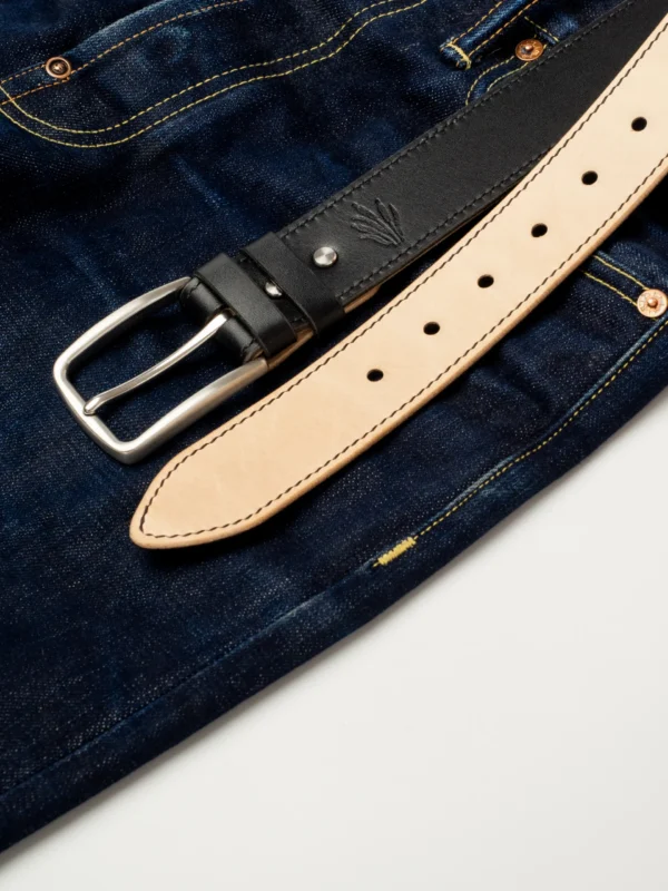 pasek-buttero-habaz-czarny-stylizacja-jeans Czarny pasek Buttero ze stalową klamrą, leżący na parze ciemnych, surowych jeansów.