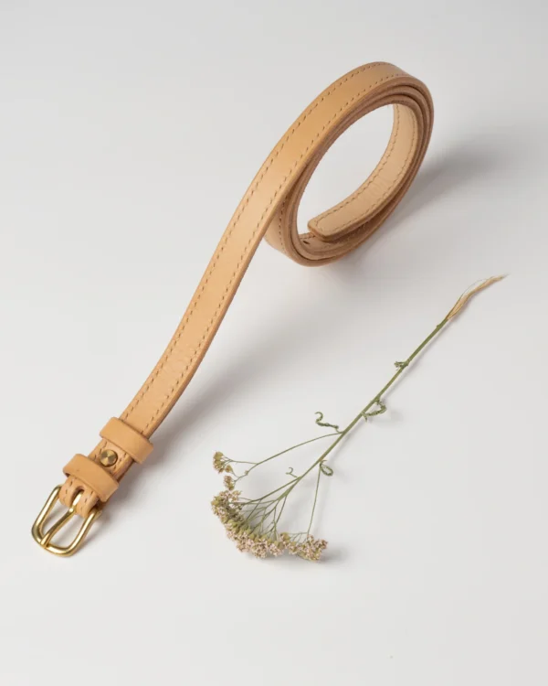 pasek-buttero-habaz-naturalny-glowny-pojedynczy Skórzany pasek Buttero w kolorze naturalnym (jasny beż), zwinięty w spiralę, z mosiężną klamrą, obok suchy polny kwiatek.