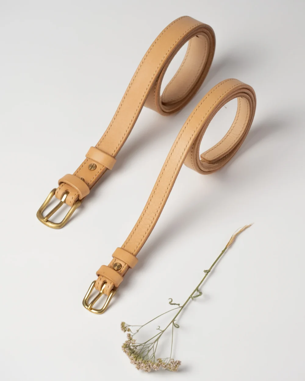 pasek-buttero-habaz-naturalny-produkt-dwa Dwa skórzane paski Buttero w kolorze naturalnym, z mosiężnymi klamrami, leżące równolegle.