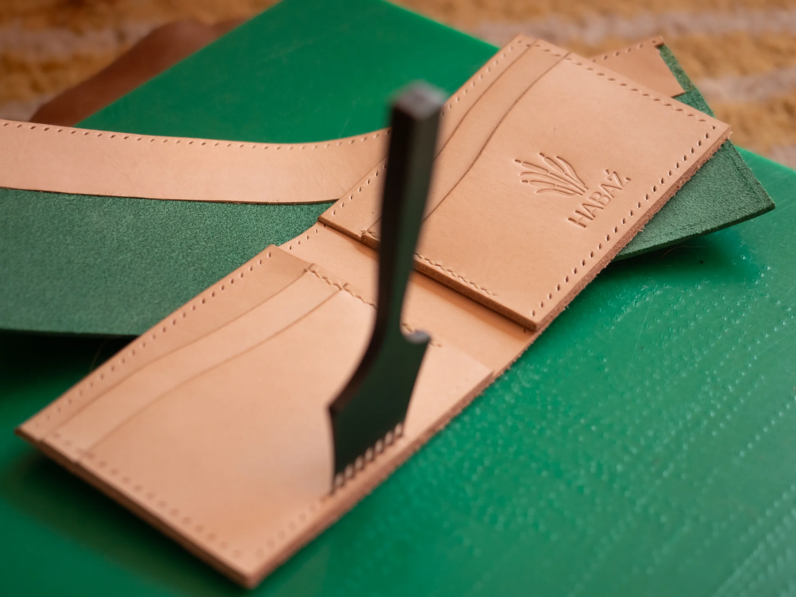 Otwarty, skórzany portfel w kolorze naturalnym, z wytłoczonym logo Habaź, leżący na zielonej podkładce, widoczny sprzęt do wykańczania krawędzi.