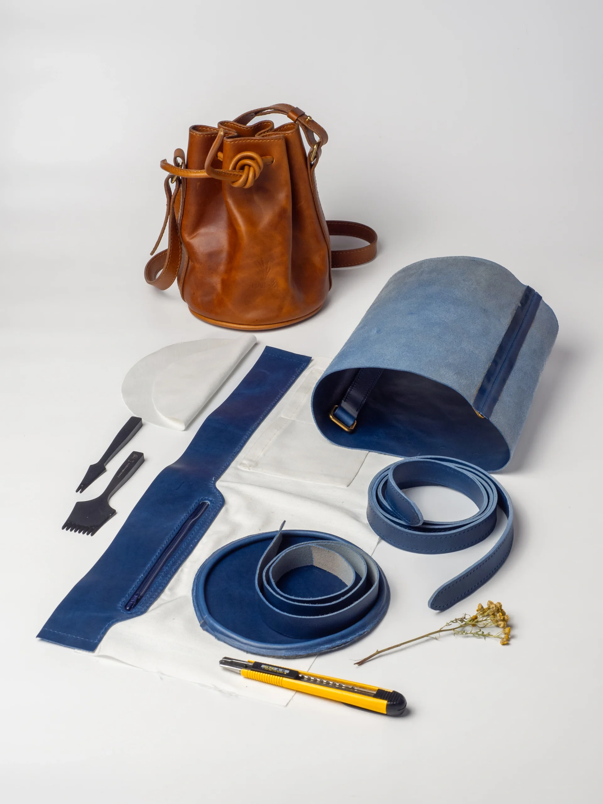Składniki do produkcji torebek: gotowa karmelowa torebka-worek, obok wycięte elementy torebki z niebieskiego zamszu i granatowej skóry licowej, nożyk, narzędzia kaletnicze i zwinięty pasek.
