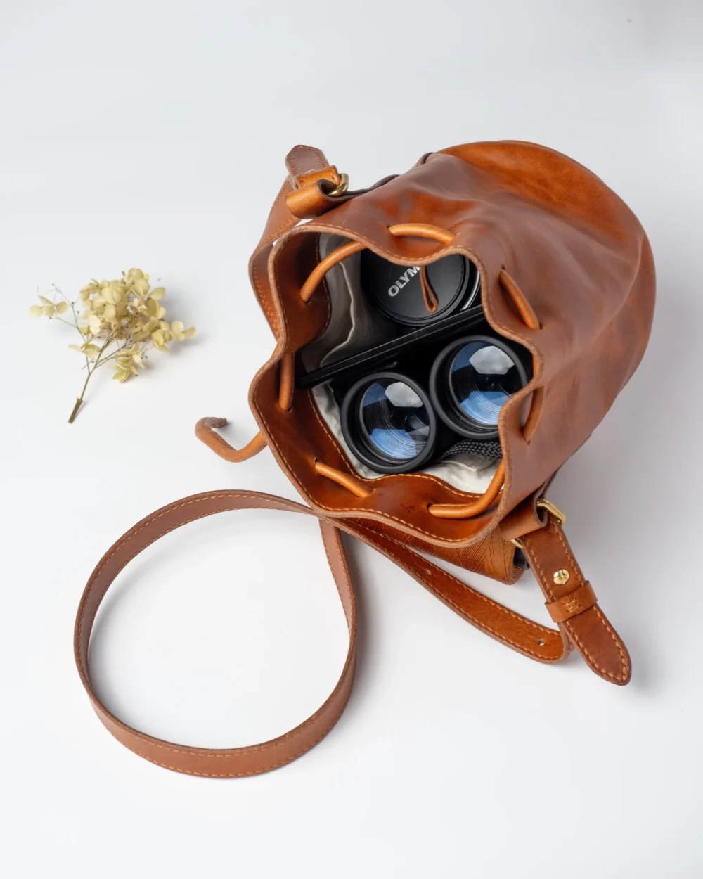 Karmelowa torebka-worek z lornetką i obiektywami w środku, otoczona suchymi kwiatami.
