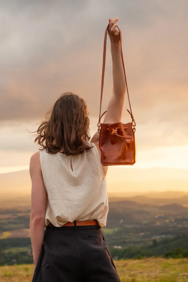 torebka-nr3-habaz-karmelowa-stylizacja-podniesiona-ręka-zachód Kobieta podnosi karmelową torebkę-worek na tle zachodzącego słońca w plenerze.