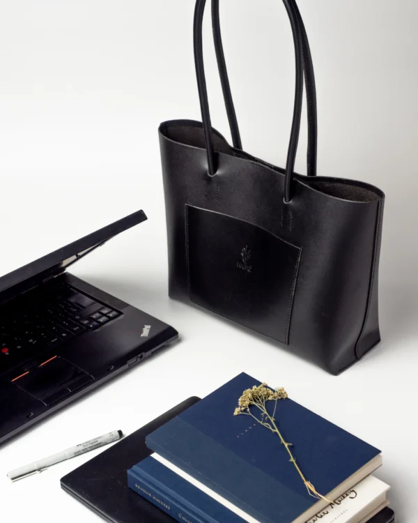 torebka-nr4-habaz-czarny-biuro-laptop Czarna torebka Shopper obok otwartego, czarnego laptopa i stosu notesów na białym biurku.