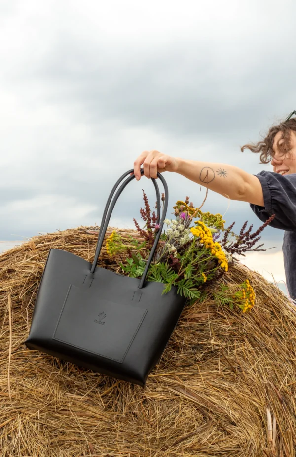 torebka-nr4-habaz-czarny-stylizacja-kwiaty Czarna skórzana torebka typu shopper (tote bag), wypełniona bukietem polnych kwiatów, trzymana przez osobę stojącą na stogu siana.