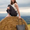 torebka-nr4-habaz-czarny-stylizacja-modelka Modelka siedząca boso na stogu siana na tle górskiego krajobrazu, obok niej leży czarna skórzana torebka Shopper.