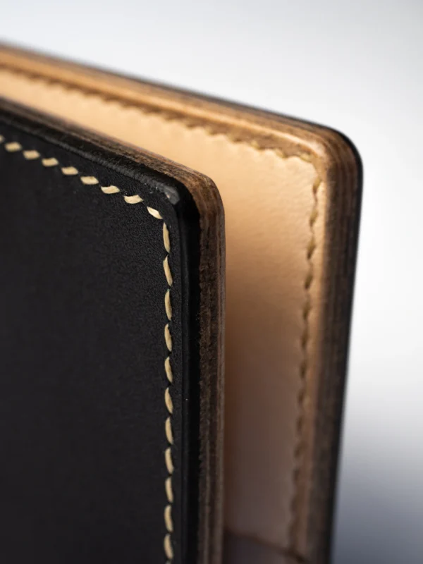 portfel-habaz-czarny-zbliżenie-krawędź-kontrast Zbliżenie na róg czarnego portfela Habaź, ukazujące kontrastowe beżowe szycie i dwie warstwy skóry: czarną zewnętrzną i jasną wewnętrzną.