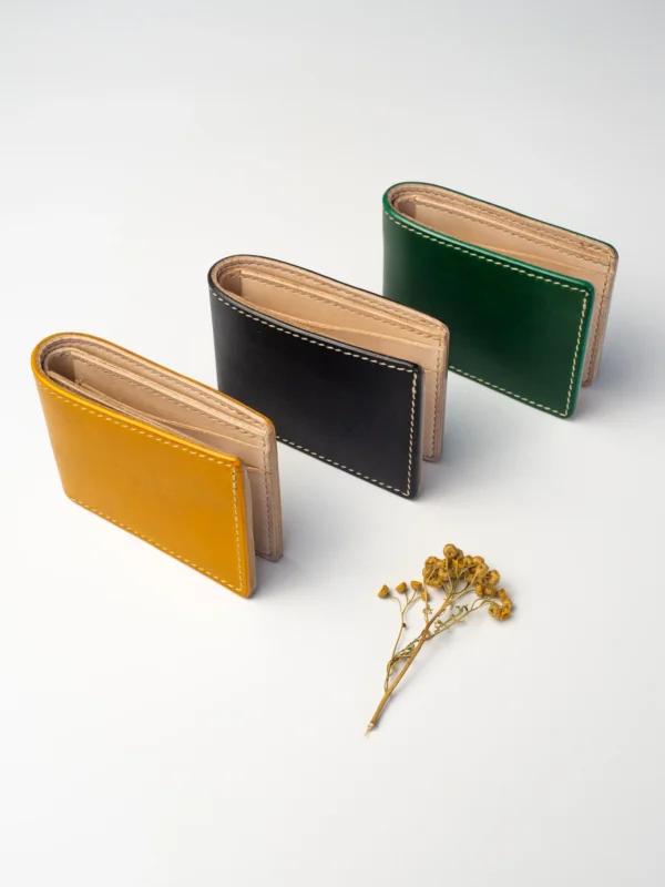 Trzy portfele Habaź (musztardowy, czarny, zielony) ustawione w rzędzie, z beżowym wnętrzem skierowanym do góry, na białym tle z suszonymi kwiatami.