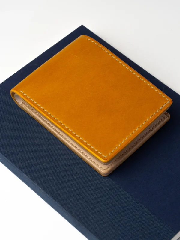 portfel-habaz-musztardowy-góra-na-notatniku Musztardowy, skórzany portfel Habaź położony na granatowym notatniku, ujęcie z góry. Widoczne beżowe szycie i jasne wnętrze portfela.