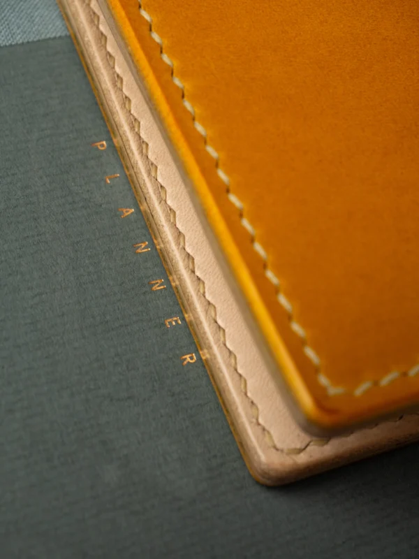 portfel-habaz-musztardowy-zbliżenie-krawędź-planner Ekstremalne zbliżenie na krawędź musztardowego portfela Habaź, spoczywającego na plannerze ze złotym napisem. Widoczne warstwy skóry i beżowe szycie.