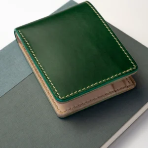 Kompaktowy, ciemnozielony portfel Habaź położony na dwóch notatnikach (zielonym i niebieskim), z widocznym beżowym, kontrastowym szwem.