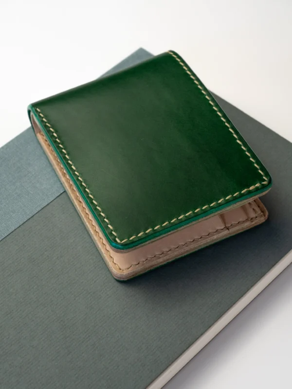portfel-habaz-zielony-góra-na-notatniku Kompaktowy, ciemnozielony portfel Habaź położony na dwóch notatnikach (zielonym i niebieskim), z widocznym beżowym, kontrastowym szwem.