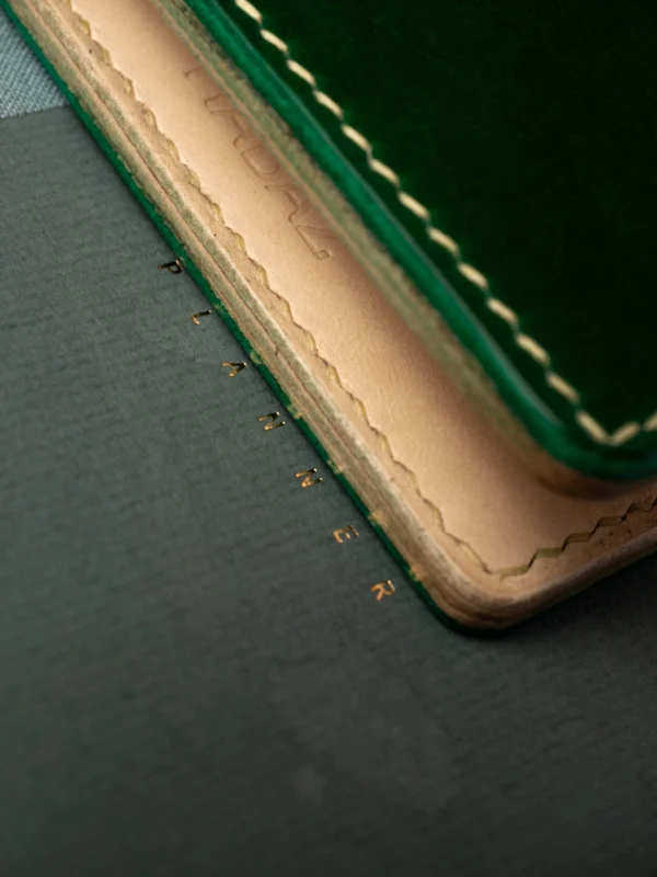 Ekstremalne zbliżenie na krawędź zielonego portfela Habaź, spoczywającego na planerze ze złotym napisem. Widoczne szczegóły barwienia krawędzi i surowa skóra wnętrza.