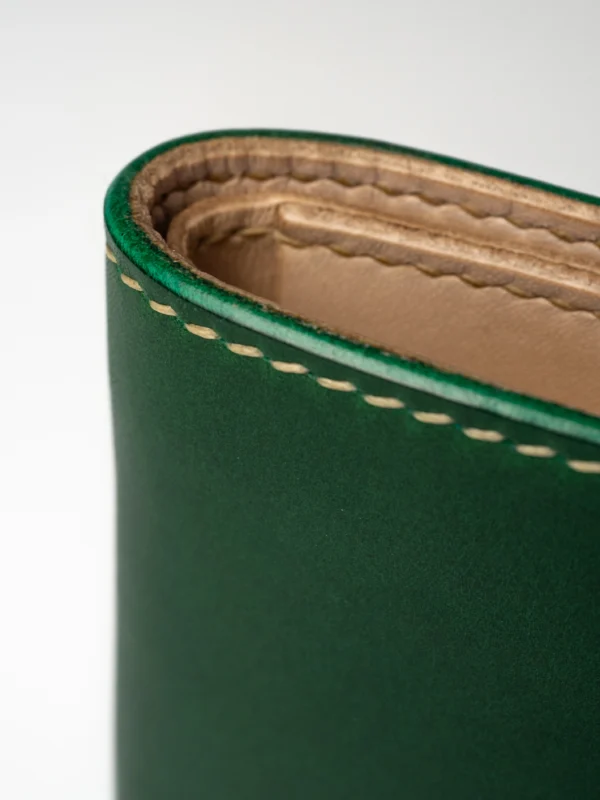 portfel-habaz-zielony-zbliżenie-rant-szycie Zbliżenie na zaokrąglony róg zielonego skórzanego portfela Habaź, ukazujące dokładne beżowe szycie i ręcznie wykończone, barwione na zielono krawędzie.